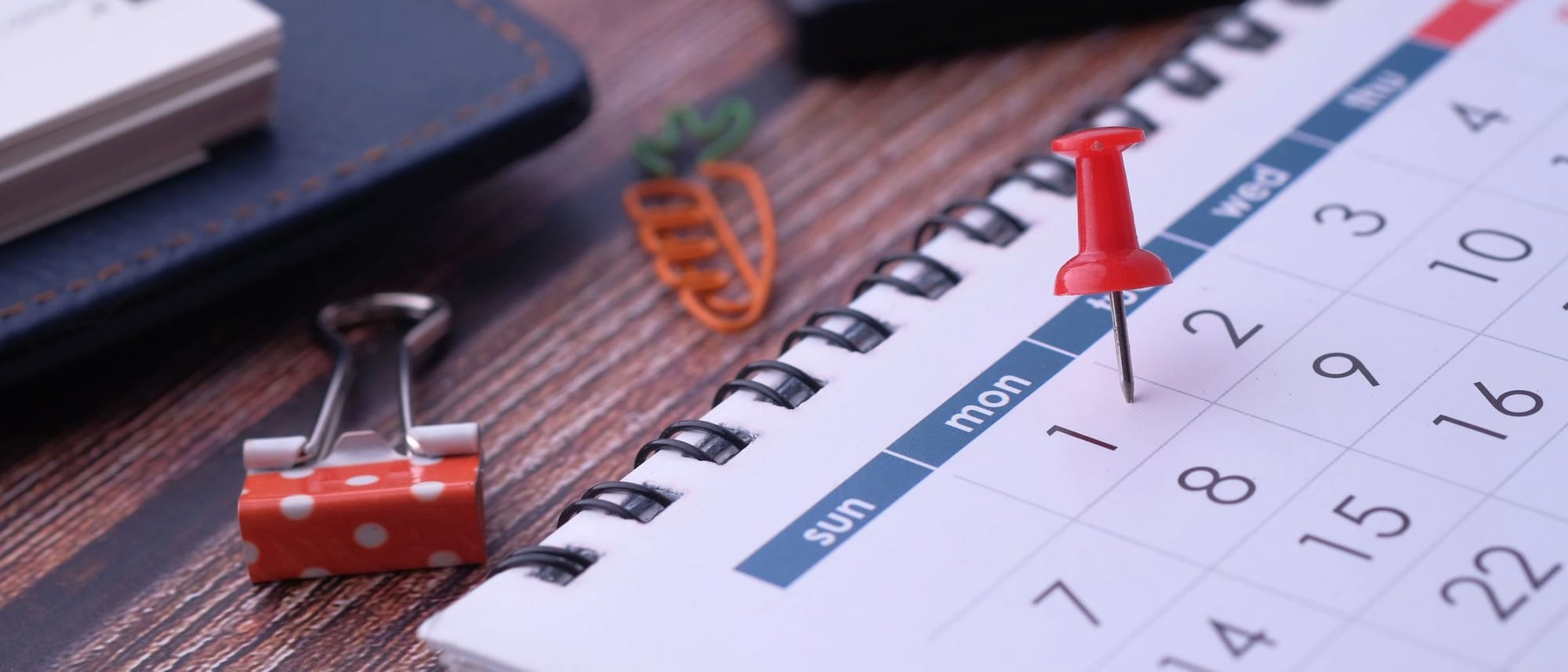 Rote Pinnnadel auf einem Tischkalender, daneben Kugelschreiber und Papierklammer auf einem Holztisch.