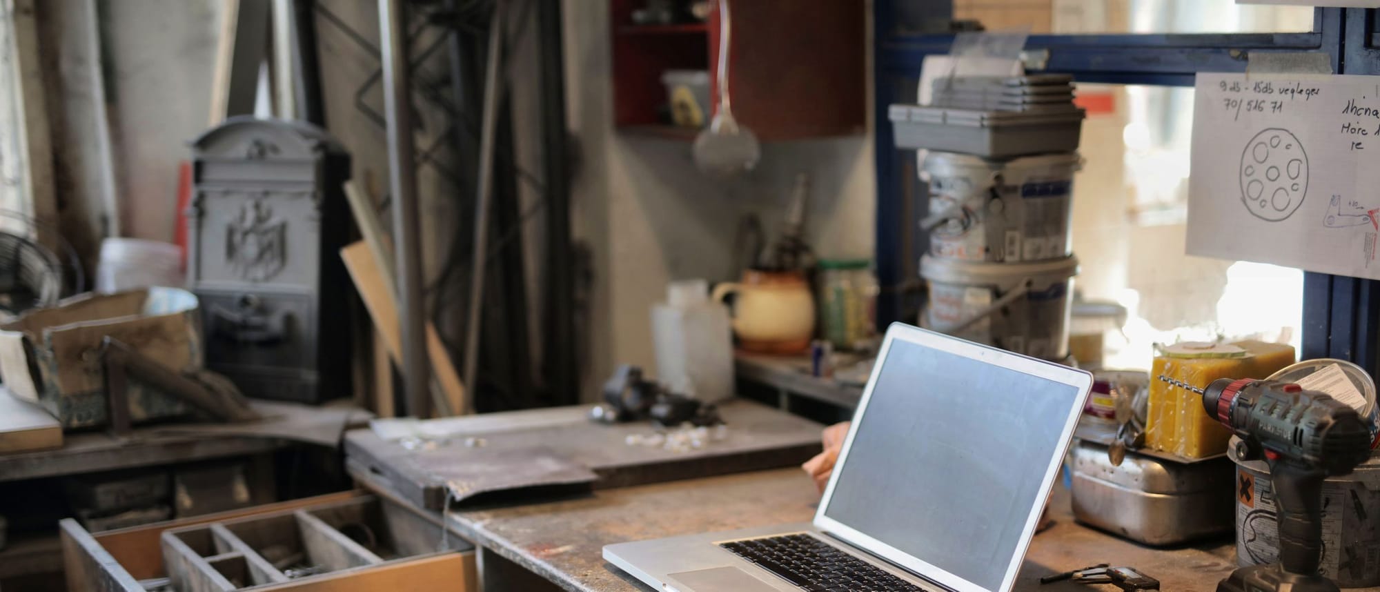 Aufgeklappter Laptop auf einer Werkbank zwischen Werkzeug, Farbeimern und offenen Schubladen.