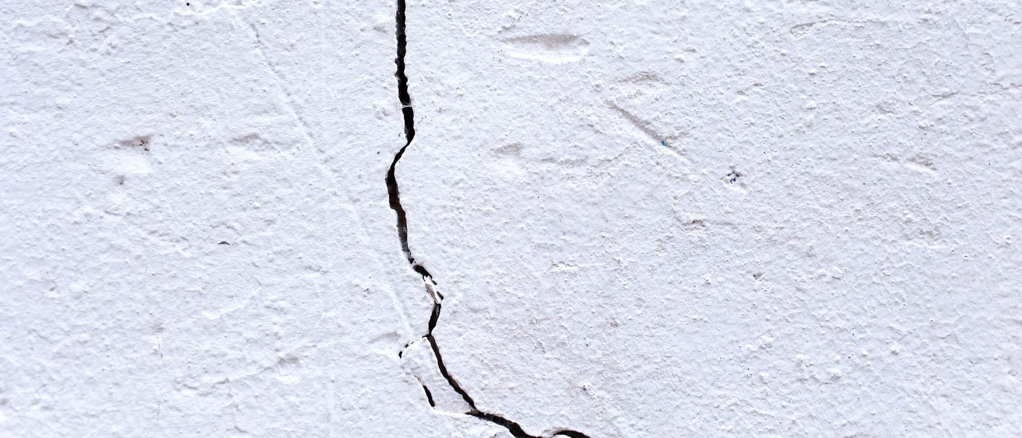 Senkrechter, dunkler Riss in einer weiß gestrichenen, rau verputzten Wand, aus der Nähe fotografiert.