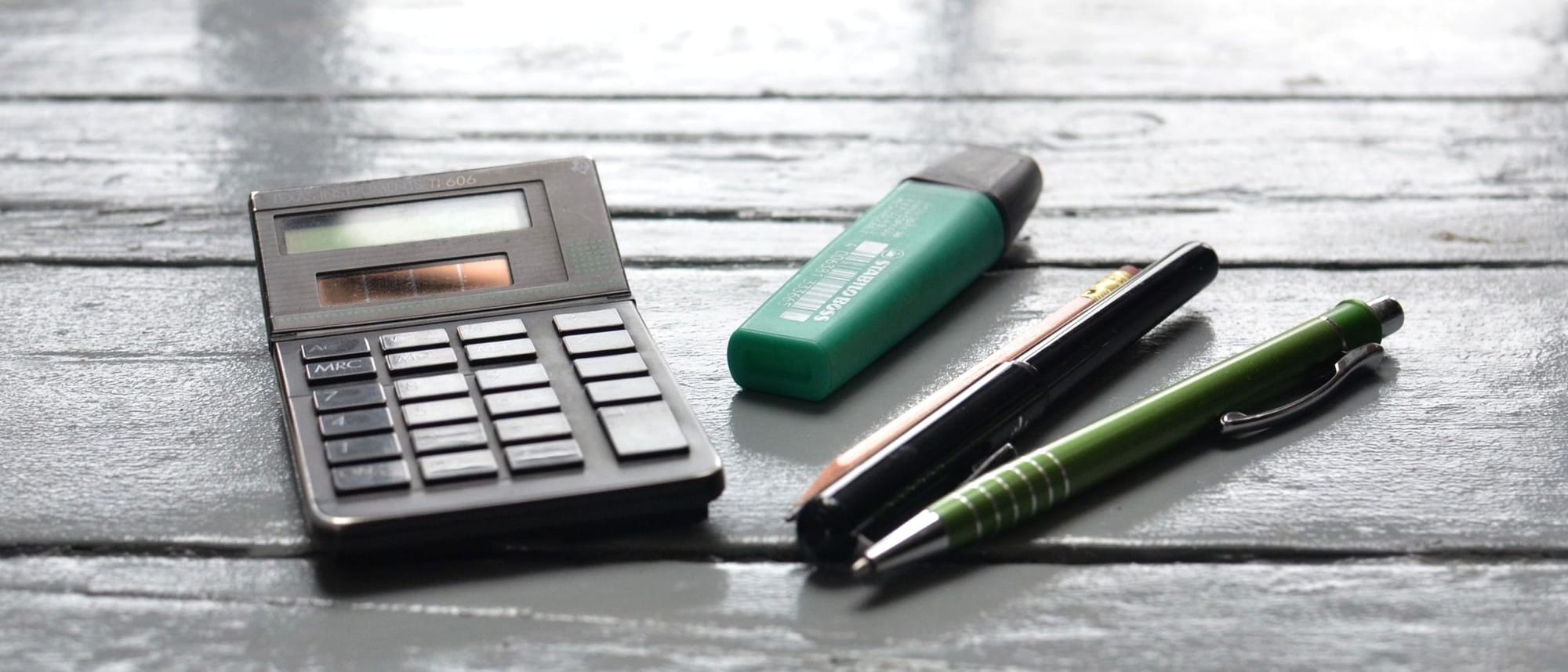 Taschenrechner, Textmarker und zwei Stifte auf dunkel gebeiztem Holz.