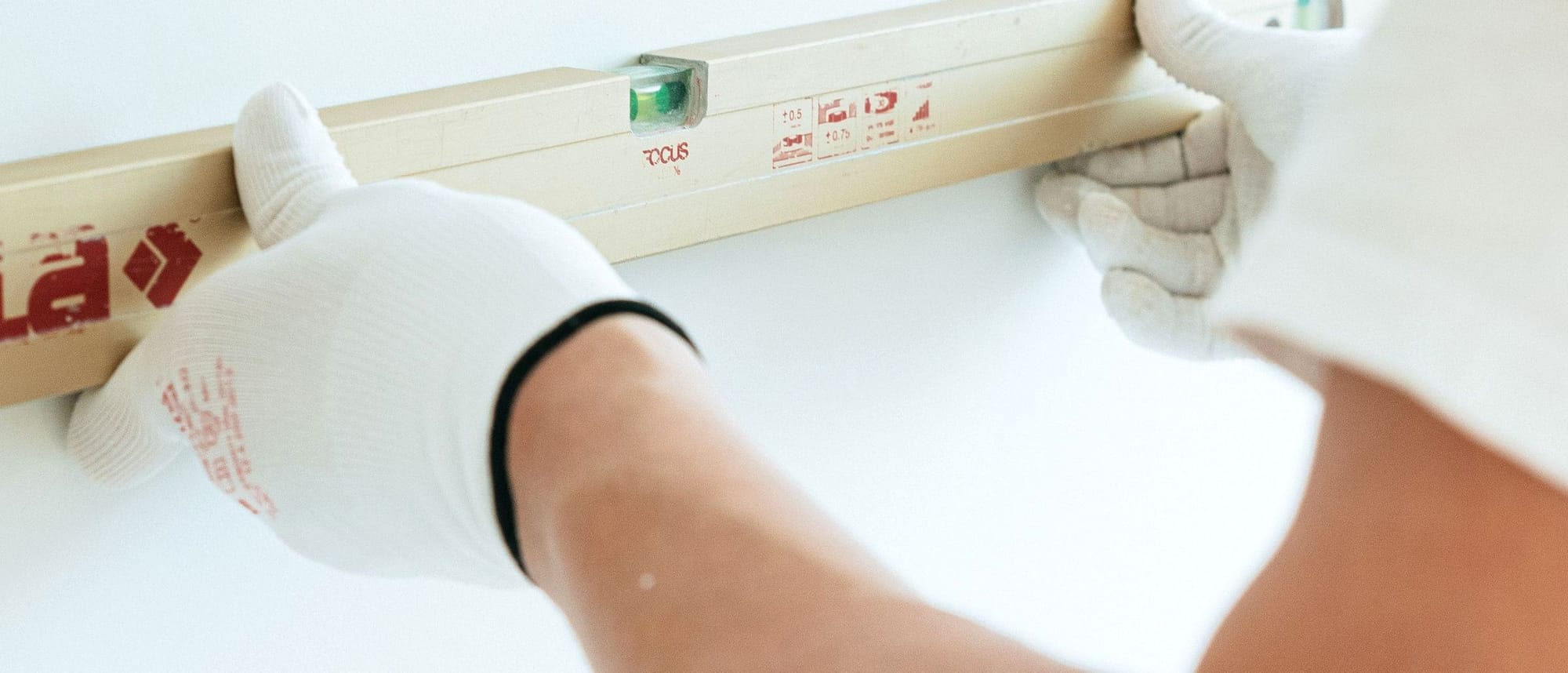 Hände in Arbeitshandschuhen halten eine Wasserwaage an eine helle, fertige Wand.