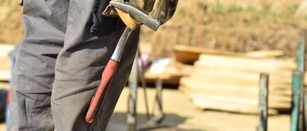 Arbeitshose mit Werkzeuggurt und Hammer auf der Baustelle, dahinter gestapelte Bretter.