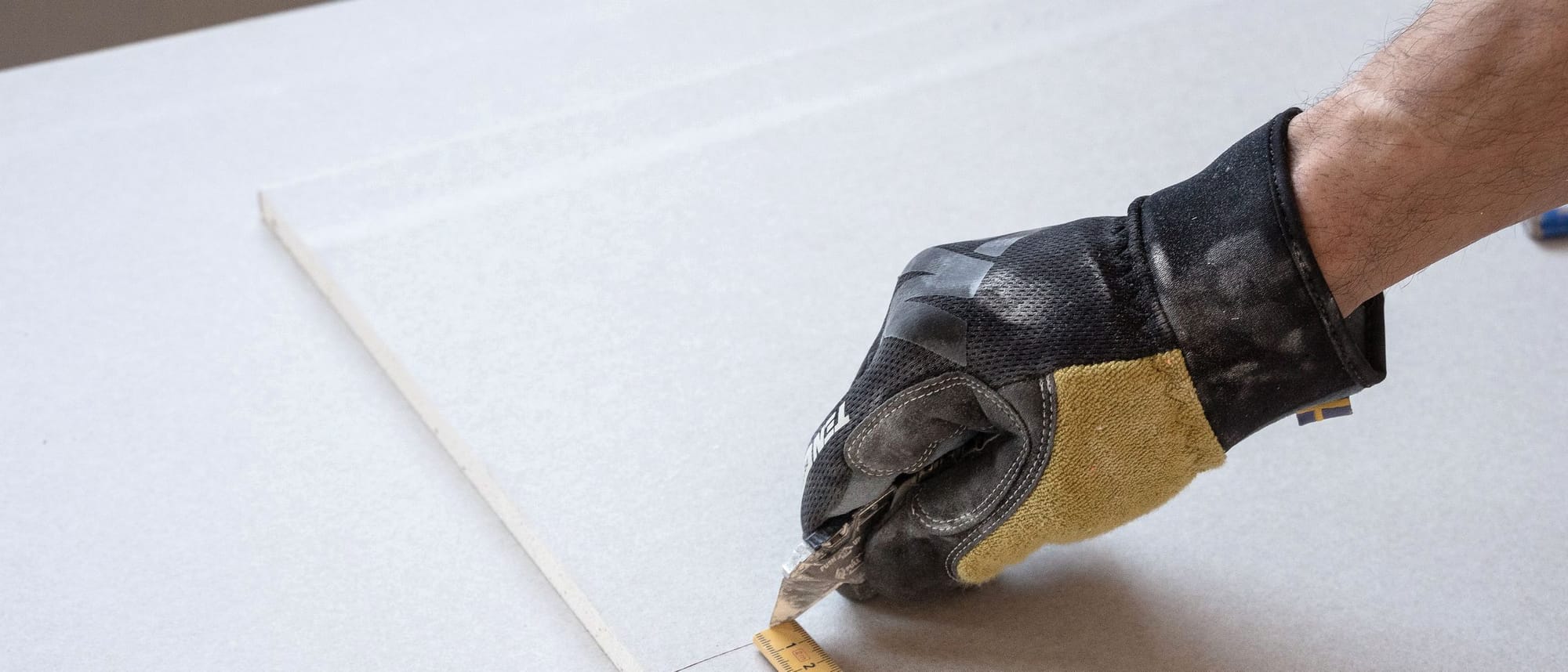Eine Hand im Arbeitshandschuh markiert mit einem Zollstock ein Maß auf einer Gipskartonplatte.